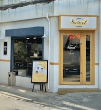 A small corner cafe with a modern facade. The entrance features natural wood-framed glass doors with a 'Marcel' sign above. A sidewalk poster advertises limited seating of 10. Inside, there is visible greenery and minimalist seating.
