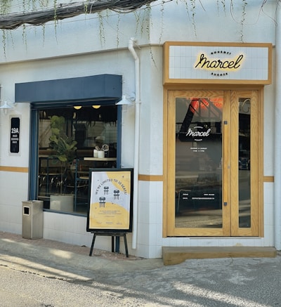 A small corner cafe with a modern facade. The entrance features natural wood-framed glass doors with a 'Marcel' sign above. A sidewalk poster advertises limited seating of 10. Inside, there is visible greenery and minimalist seating.