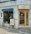 A small corner cafe with a modern facade. The entrance features natural wood-framed glass doors with a 'Marcel' sign above. A sidewalk poster advertises limited seating of 10. Inside, there is visible greenery and minimalist seating.