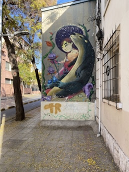 A street mural features a whimsical scene with a young person sitting next to a large, friendly otter holding a cup. Colorful lilies and decorative leaves surround them, and the person appears to be nestled comfortably with a look of contentment on their face. The mural is located on the side of a building, partially framed by the urban environment including a tree and sidewalk.