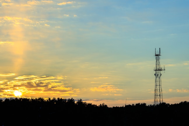 A telecommunications tower stands prominently against a serene sky, partially illuminated by the warm hues of a setting sun. The horizon is lined with dark silhouettes of trees, and soft clouds are lit with golden and orange shades, creating a peaceful contrast with the sky's calming blues.