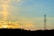 A telecommunications tower stands prominently against a serene sky, partially illuminated by the warm hues of a setting sun. The horizon is lined with dark silhouettes of trees, and soft clouds are lit with golden and orange shades, creating a peaceful contrast with the sky's calming blues.