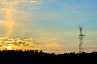 A telecommunications tower stands prominently against a serene sky, partially illuminated by the warm hues of a setting sun. The horizon is lined with dark silhouettes of trees, and soft clouds are lit with golden and orange shades, creating a peaceful contrast with the sky's calming blues.