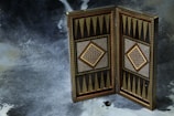 An intricately patterned backgammon board is displayed in an upright position, showcasing geometric designs and detailed ornamentation. The surface beneath the board is textured and appears to be fur-like. A pair of dice and a black game piece are placed in front of the board, contributing to the backgammon theme.