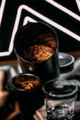 A close-up view of coffee beans inside a metal container, with additional beans in a smaller open jar next to it. A clear glass cup is situated in the foreground, and a patterned black backdrop with white lines adds contrast.