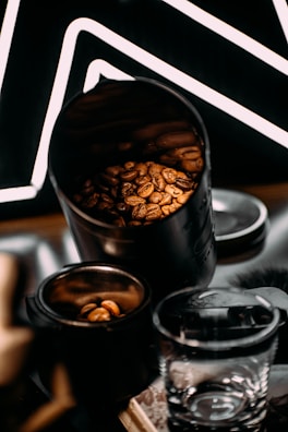 A close-up view of coffee beans inside a metal container, with additional beans in a smaller open jar next to it. A clear glass cup is situated in the foreground, and a patterned black backdrop with white lines adds contrast.