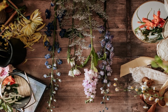 A wooden surface is adorned with various floral elements, including blue, purple, white, and pink flowers arranged intricately. The scene includes a few reflective surfaces like mirrors that capture parts of the flowers and the surroundings. A black vase holds some yellow petals, and there's a book or notebook partly visible. Fabrics and a spool of thread add texture to the tableau, conveying a sense of creativity.