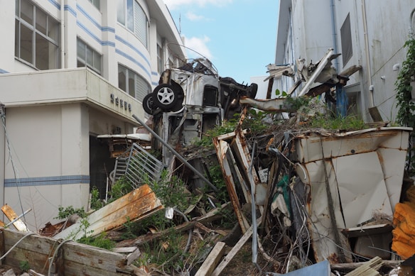 A scene of destruction and debris featuring numerous wrecked objects, including an overturned vehicle lying amidst twisted metal, broken wood, and felled vegetation. The area is surrounded by damaged buildings with visible structural impacts. There is considerable overgrowth and rust on the metal parts.
