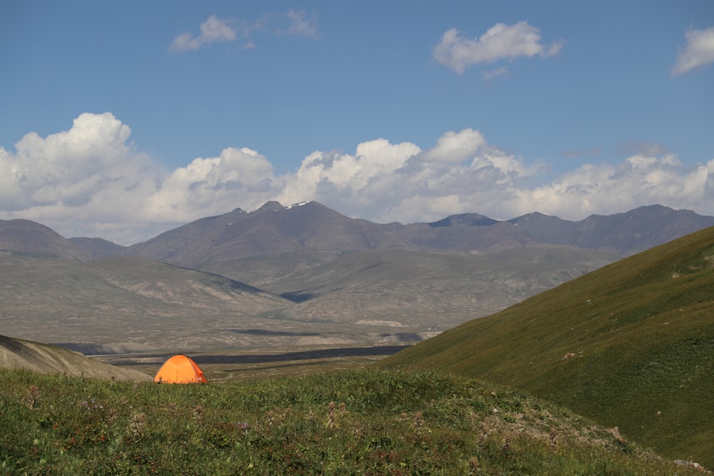 orange tent on green grass field near mountains under blue and white sunny cloudy sky during