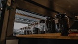Several glass jars filled with dark coffee beans are placed on a wooden shelf. The setting appears to be indoors with a metallic roof visible through an opening, suggesting a rustic or industrial ambiance.