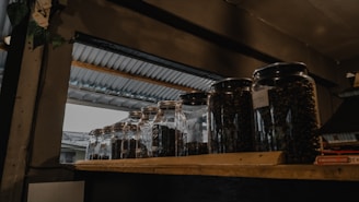 Several glass jars filled with dark coffee beans are placed on a wooden shelf. The setting appears to be indoors with a metallic roof visible through an opening, suggesting a rustic or industrial ambiance.