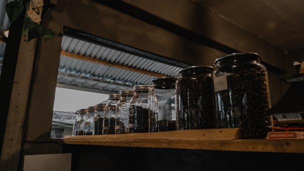 Several glass jars filled with dark coffee beans are placed on a wooden shelf. The setting appears to be indoors with a metallic roof visible through an opening, suggesting a rustic or industrial ambiance.
