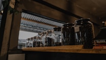 Several glass jars filled with dark coffee beans are placed on a wooden shelf. The setting appears to be indoors with a metallic roof visible through an opening, suggesting a rustic or industrial ambiance.