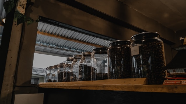 Several glass jars filled with dark coffee beans are placed on a wooden shelf. The setting appears to be indoors with a metallic roof visible through an opening, suggesting a rustic or industrial ambiance.