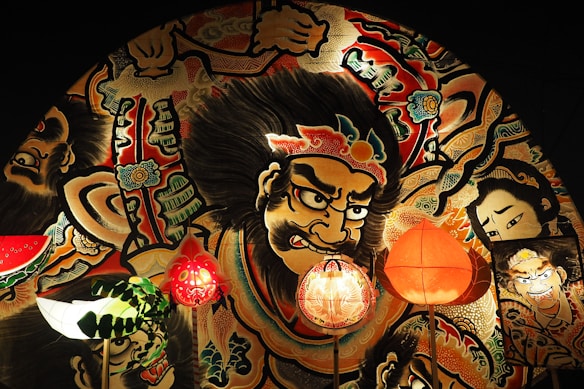 A vibrant and detailed mural with powerful, expressive characters painted in an intricate style. The central figure features a fierce expression, adorned with decorative patterns and motifs that suggest a traditional or cultural theme. A variety of colorful lanterns are positioned in front of the mural, adding depth and warmth.