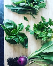 A wooden cutting board is adorned with fresh herbs and a small purple root vegetable with attached greens. The herbs, including basil, parsley, and arugula, are arranged neatly with visible vibrant green leaves. A small pile of dark soil accompanies the root vegetable, suggesting freshness and organic growth.