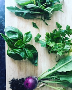 A wooden cutting board is adorned with fresh herbs and a small purple root vegetable with attached greens. The herbs, including basil, parsley, and arugula, are arranged neatly with visible vibrant green leaves. A small pile of dark soil accompanies the root vegetable, suggesting freshness and organic growth.