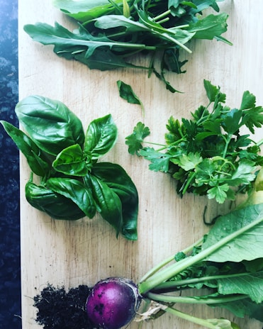 A wooden cutting board is adorned with fresh herbs and a small purple root vegetable with attached greens. The herbs, including basil, parsley, and arugula, are arranged neatly with visible vibrant green leaves. A small pile of dark soil accompanies the root vegetable, suggesting freshness and organic growth.