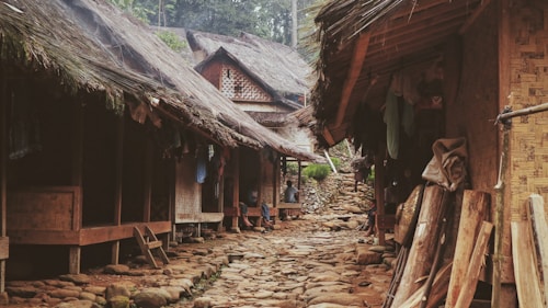Traditional wooden huts with thatched roofs line a narrow, stone-paved village path. The rustic architecture features woven bamboo walls and wooden supports, with a variety of natural materials visible. Lush greenery surrounds the area, adding to the serene and earthy atmosphere.