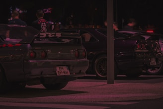 Two parked sports cars are visible under low light conditions, featuring aftermarket modifications and decals. The background shows people conversing near the vehicles in a nightlife setting.