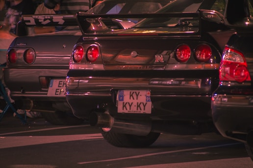 Several cars are parked closely together, with a focus on the rear view of two Nissan Skyline GT-R models. The cars have distinctive round taillights and customized license plates. The scene appears to be at a car meet, surrounded by people and illuminated with a warm, ambient light.