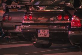 Several cars are parked closely together, with a focus on the rear view of two Nissan Skyline GT-R models. The cars have distinctive round taillights and customized license plates. The scene appears to be at a car meet, surrounded by people and illuminated with a warm, ambient light.