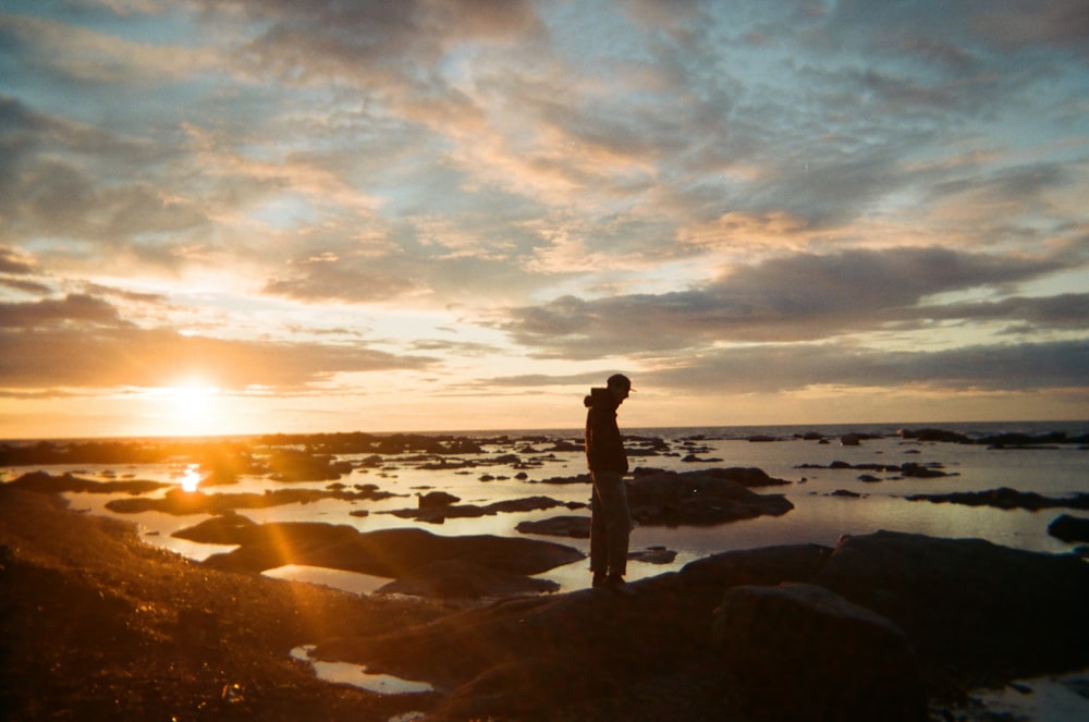 silhouette of person standing on rock formation near body of water during sunset