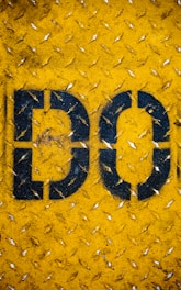 A close-up view of a yellow textured metal surface, featuring the bold black letters 'DO' painted on it. The surface appears to be weathered, with scratches and small rust spots visible.