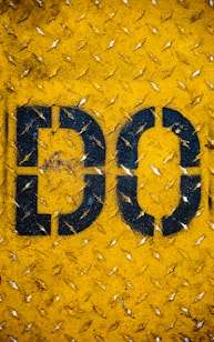 A close-up view of a yellow textured metal surface, featuring the bold black letters 'DO' painted on it. The surface appears to be weathered, with scratches and small rust spots visible.