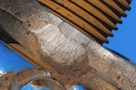 A close-up view of a textured metallic surface with visible wear and rust marks. The image includes various shades of metal, showing both shiny and tarnished areas. In the background, parts of a blue structure and ridged orange component are visible.