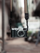 A vintage-style camera with a textured body and metallic elements is suspended by a colorful, woven strap. The background appears blurred, hinting at an outdoor urban setting, enhancing the camera's prominence.