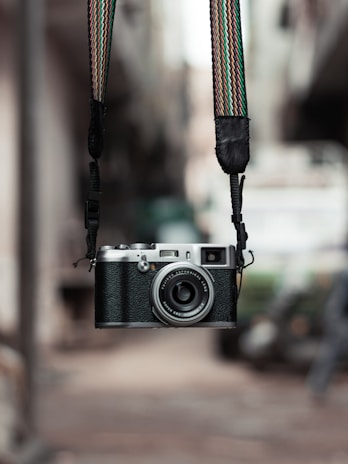 A vintage-style camera with a textured body and metallic elements is suspended by a colorful, woven strap. The background appears blurred, hinting at an outdoor urban setting, enhancing the camera's prominence.