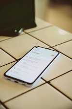 A smartphone is placed on a white tiled surface. The phone screen displays the settings menu, featuring options such as Network & Internet and Connected devices. The lighting is soft, casting a slight shadow to the left of the phone.