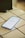A smartphone is placed on a white tiled surface. The phone screen displays the settings menu, featuring options such as Network & Internet and Connected devices. The lighting is soft, casting a slight shadow to the left of the phone.