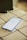 A smartphone is placed on a white tiled surface. The phone screen displays the settings menu, featuring options such as Network & Internet and Connected devices. The lighting is soft, casting a slight shadow to the left of the phone.