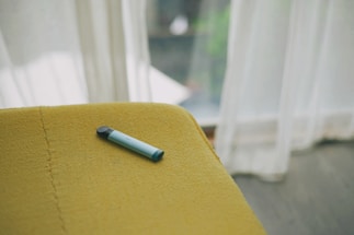 A vape pen with a black and light blue color scheme rests on a mustard yellow upholstered surface. Natural light streams through sheer white curtains in the background, creating a soft, inviting ambiance.