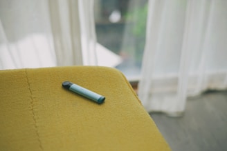 A vape pen with a black and light blue color scheme rests on a mustard yellow upholstered surface. Natural light streams through sheer white curtains in the background, creating a soft, inviting ambiance.
