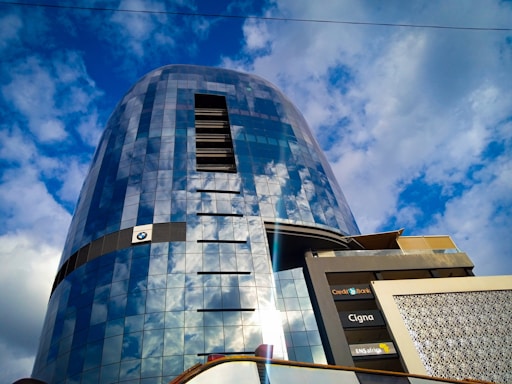 A modern, tall building with a reflective glass facade that mirrors the cloudy blue sky, creating a striking visual effect. The structure displays several brand logos, including BMW and others, near its lower part. The architectural design is sleek and contemporary, with a combination of straight lines and curved surfaces.