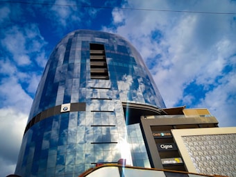 A modern, tall building with a reflective glass facade that mirrors the cloudy blue sky, creating a striking visual effect. The structure displays several brand logos, including BMW and others, near its lower part. The architectural design is sleek and contemporary, with a combination of straight lines and curved surfaces.