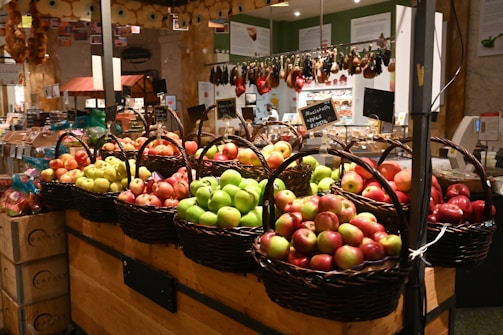 Baskets of apples in varying colors such as red, green, and yellow are displayed in a market setting. The surroundings include hanging cured meats and a variety of packaged goods, creating a bustling and vibrant atmosphere.