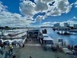 A waterfront scene with a pier extending into the water, surrounded by multiple boats docked at the piers. There are people walking around the pier area, and several bicycles are parked along a railing. High-rise buildings are visible in the background, and the sky is filled with large, fluffy clouds against a bright blue backdrop.