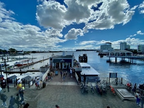 A waterfront scene with a pier extending into the water, surrounded by multiple boats docked at the piers. There are people walking around the pier area, and several bicycles are parked along a railing. High-rise buildings are visible in the background, and the sky is filled with large, fluffy clouds against a bright blue backdrop.