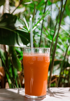 A tall glass filled with orange juice, a straw protruding from the top. It sits on a sunlit surface amidst lush green foliage, suggesting a refreshing, tropical setting.
