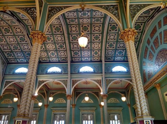Intricate architectural details featuring ornate columns with gold accents and arches supporting a patterned ceiling with star motifs. The interior is illuminated by pendant lights and natural light filtering through arched windows. The color palette includes rich teal, gold, and white tones, contributing to an opulent atmosphere.