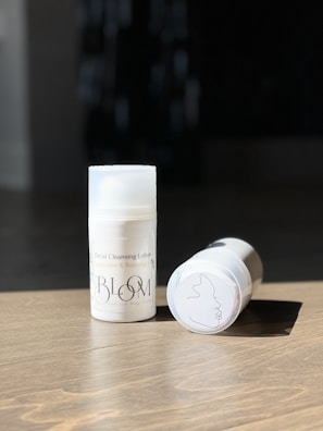 Two bottles of facial cleansing lotion labeled as Bloom sit on a wooden surface. One bottle stands upright while the other lies on its side, showing a line art drawing on its base. The background is dark, contrasting with the bright lighting on the bottles.