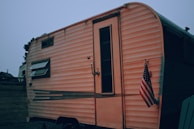 A parked vintage RV with a small American flag hanging near the door. The RV has horizontal stripes and a small window with an awning. In the background, there are faint silhouettes of other buildings or trailers.