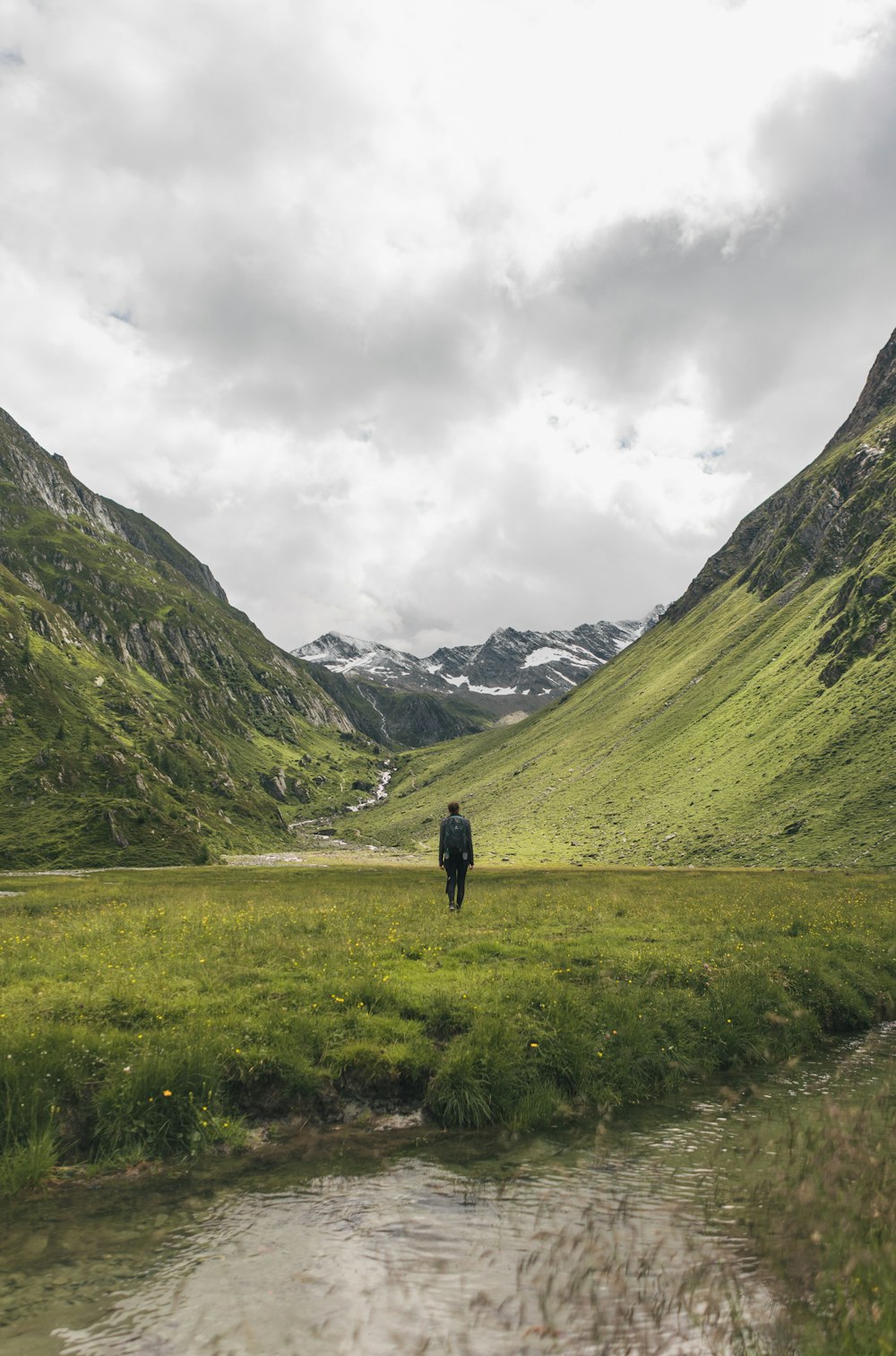 person in black jacket standing on green grass field near mountains under white cloudy sky during