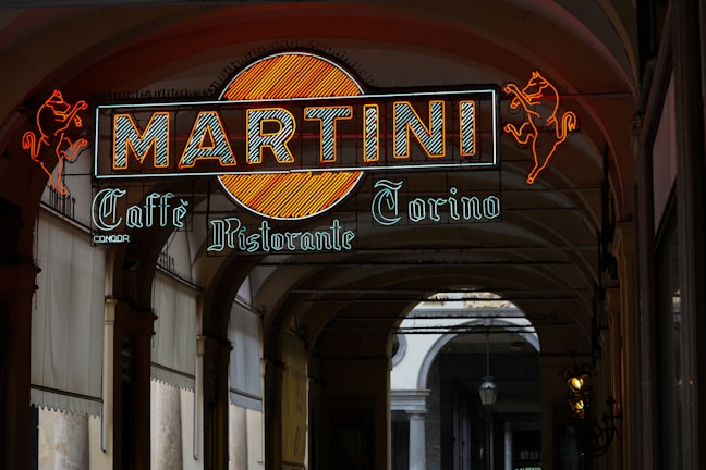 A vibrant neon sign featuring the word 'MARTINI' with a stylized orange circle behind it, accompanied by neon red horse designs on either side. The sign also includes the text 'Caffè Ristorante Torino' in a classic script style below the main title. The scene is set in an elegant arcade with archways leading to an open entrance at the far end.