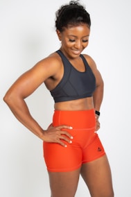 A smiling woman wearing a dark sports bra and bright orange athletic shorts with a logo on the side. She stands with her hands on her hips, exuding confidence and athleticism. Her hair is pulled back, and she seems to be in a positive mood.