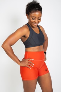 A smiling woman wearing a dark sports bra and bright orange athletic shorts with a logo on the side. She stands with her hands on her hips, exuding confidence and athleticism. Her hair is pulled back, and she seems to be in a positive mood.